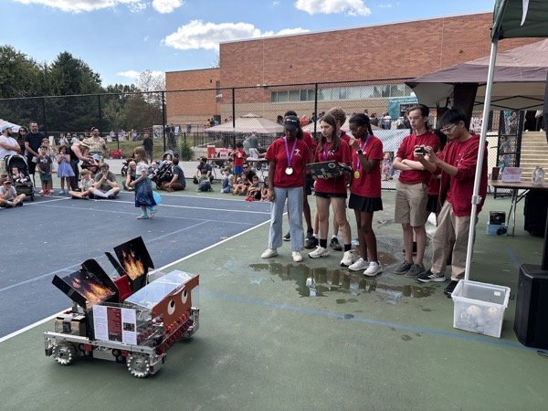 Team members wearing medals presenting a small demo robot at an outdoor community STEM event