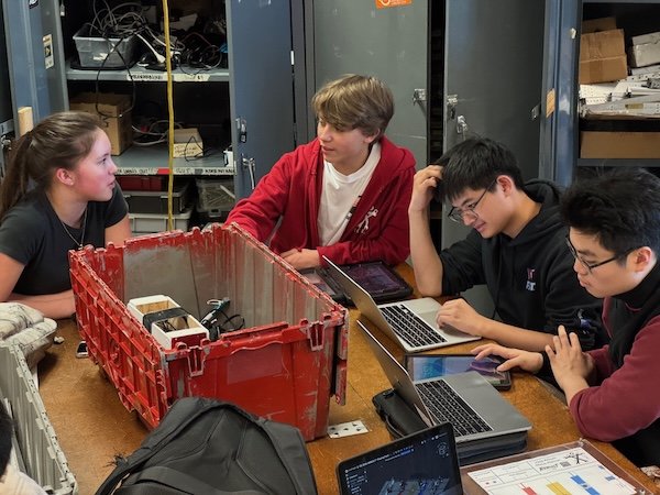 Team members collaborating on laptops around a worn parts bin during a strategy session
